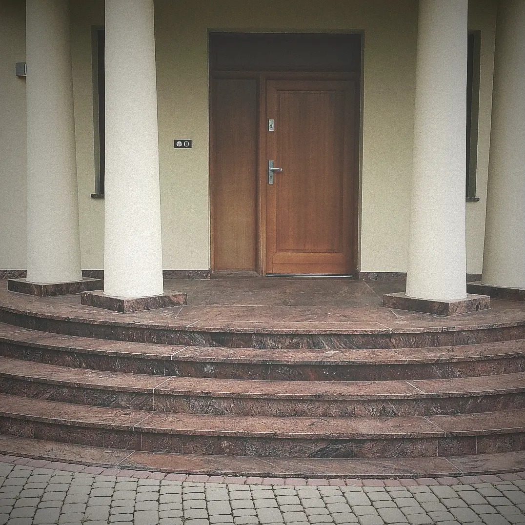 Wejście do domu z zaokrąglonymi schodami wyłożonymi brązowym kamieniem, podparte kolumnami, prowadzące do drewnianych drzwi.