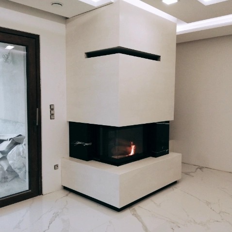 Nowoczesny, narożny kominek z białego kamienia z czarnymi wkładami szklanymi, z widocznym płomieniem, na marmurowej podłodze, w jasnym wnętrzu.
