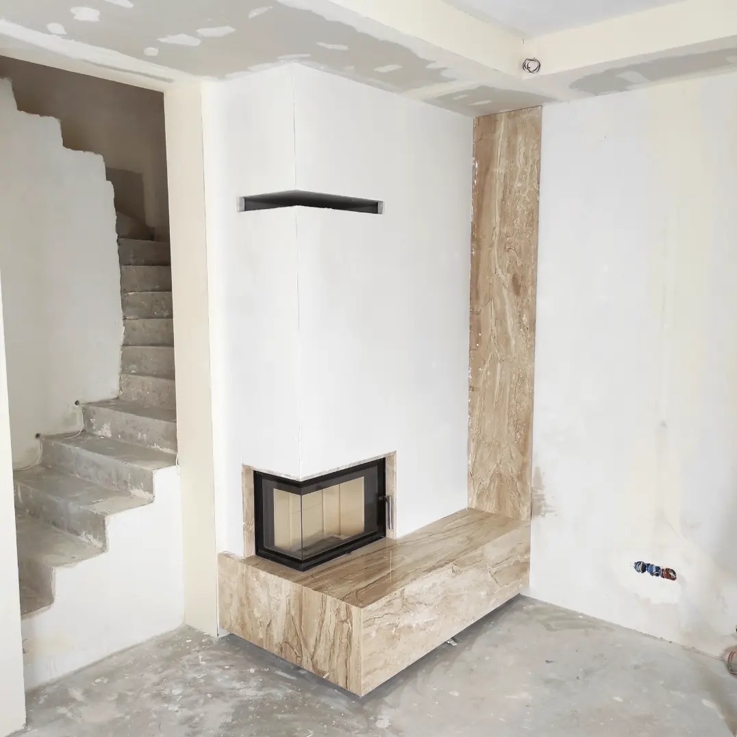 Wnętrze w trakcie wykończenia z narożnym wkładem kominkowym obudowanym płytami imitującymi marmur, widoczne surowe schody betonowe i fragment instalacji elektrycznej.