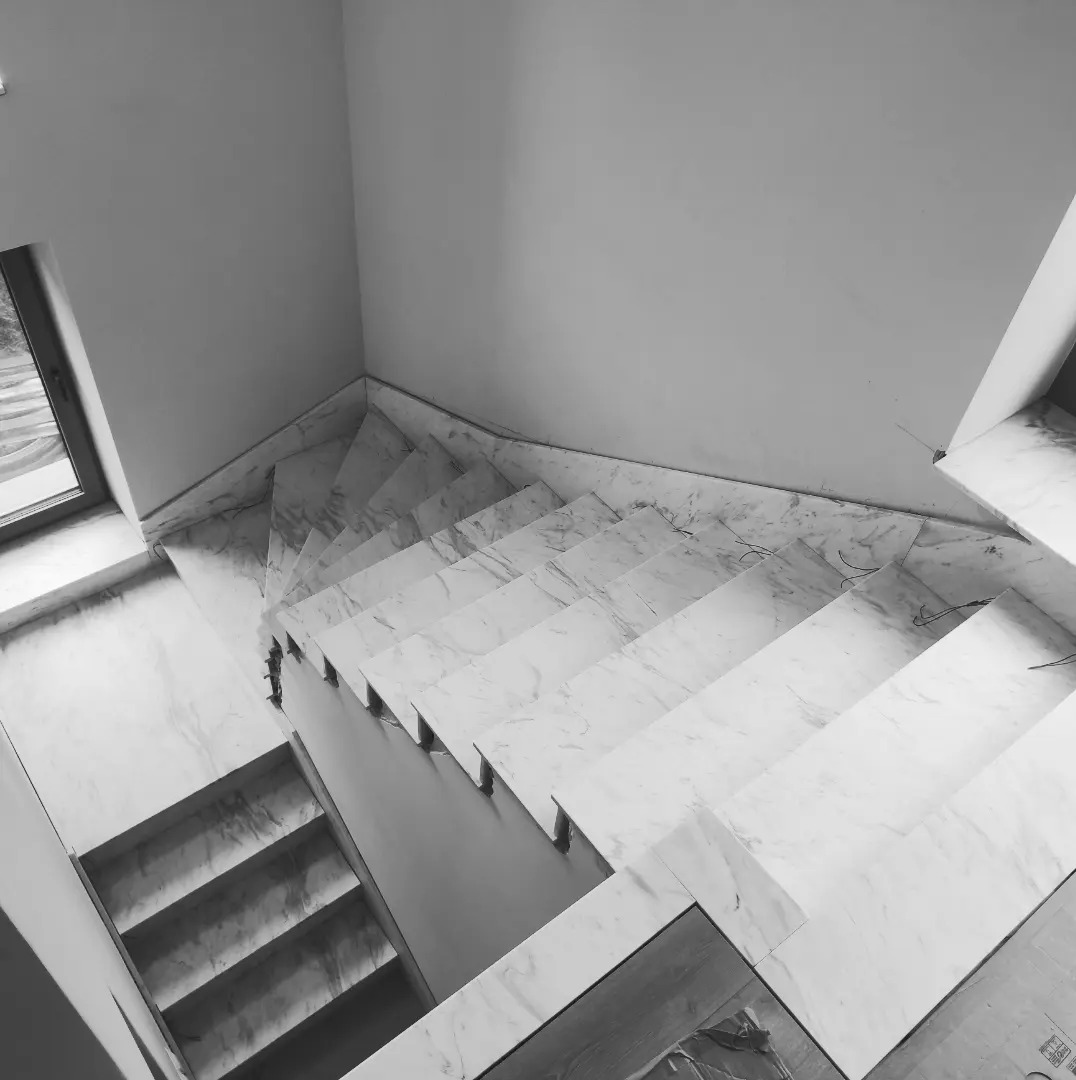 Widok z góry na spiralne schody wykończone białym marmurem, z delikatnym użyleniem, prowadzące w dół, z widocznym fragmentem okna w tle, uchwycone w czarno-białej tonacji.