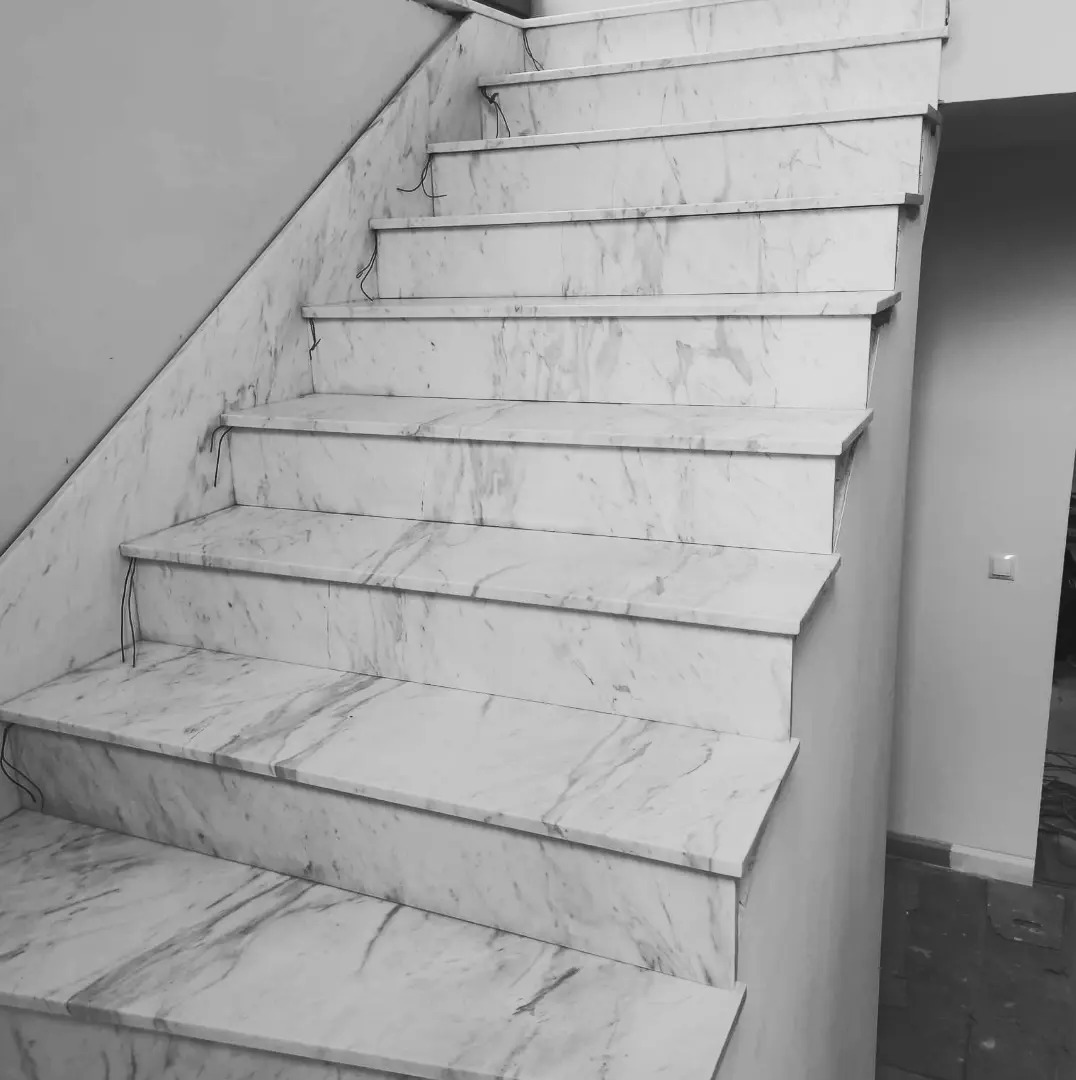 Wewnętrzne schody wykończone białym marmurem z szarymi żyłami, widoczne przewody elektryczne wystające spod stopni, monochromatyczne ujęcie.