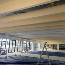 Wnętrze w trakcie malowania: jasne belki sufitowe, rusztowania i drabina. Podłoga zabezpieczona folią malarską. Duże okna z widokiem na zewnątrz.