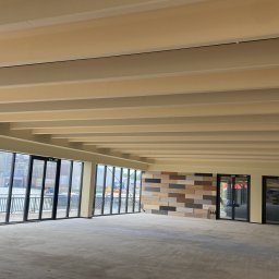 Daniel trzos - Przestronne, jasne wnętrze w trakcie wykańczania, z dużymi oknami z widokiem na wodę i dekoracyjną ścianą z paneli w odcieniach brązu i beżu. Widoczne beżowe belki stropowe.