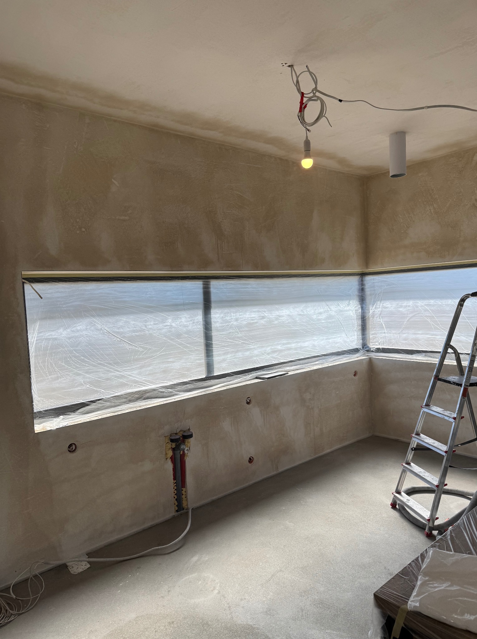 Pokój w trakcie remontu: ściany i sufit po szpachlowaniu, okno zabezpieczone folią, drabina oparta o ścianę, widoczne instalacje elektryczne i hydrauliczne.