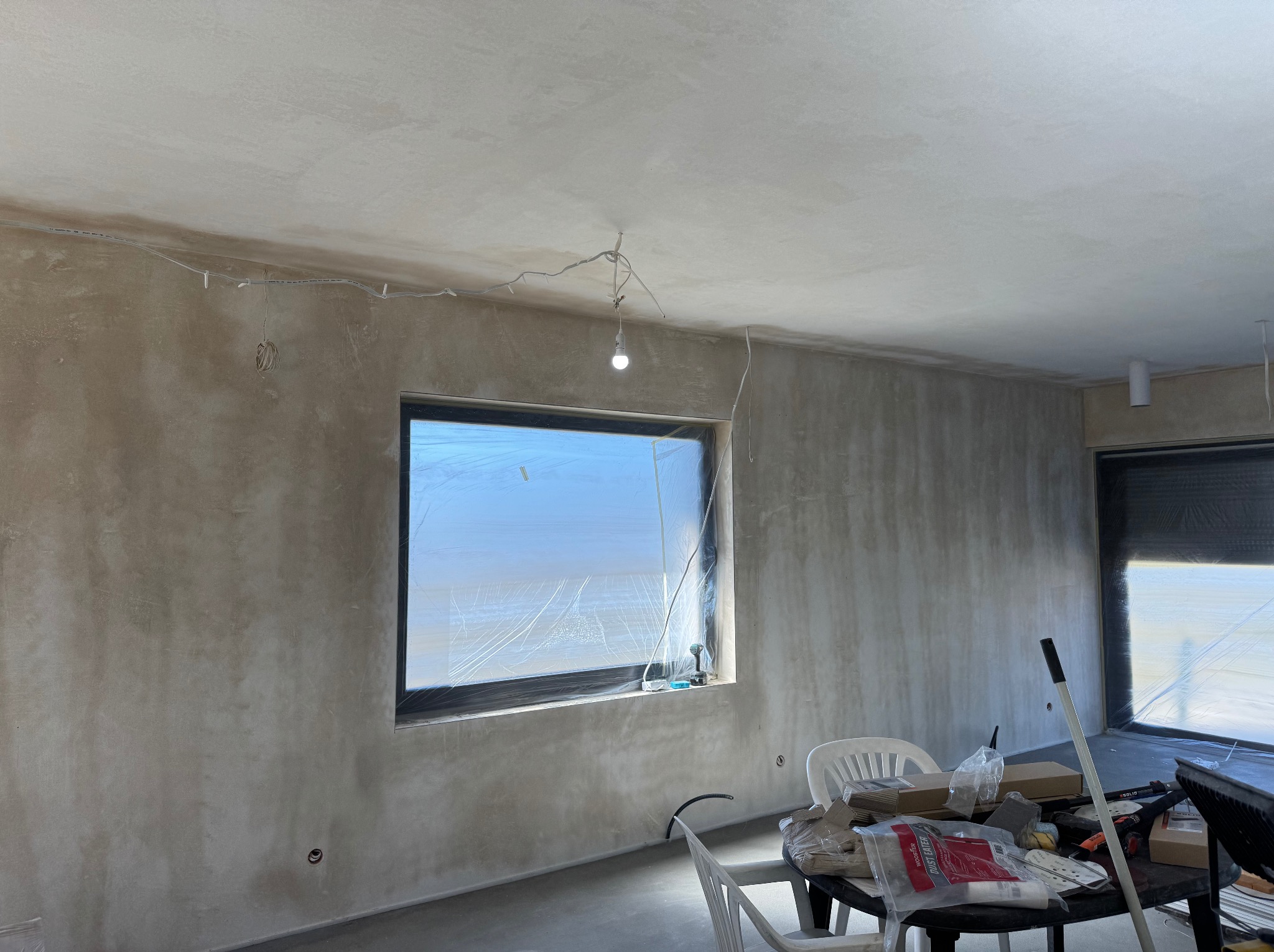 Pokój po szpachlowaniu, ściany przygotowane do malowania. Okno zabezpieczone folią, na stole narzędzia i materiały budowlane. Widoczne niedoskonałości faktury ścian.
