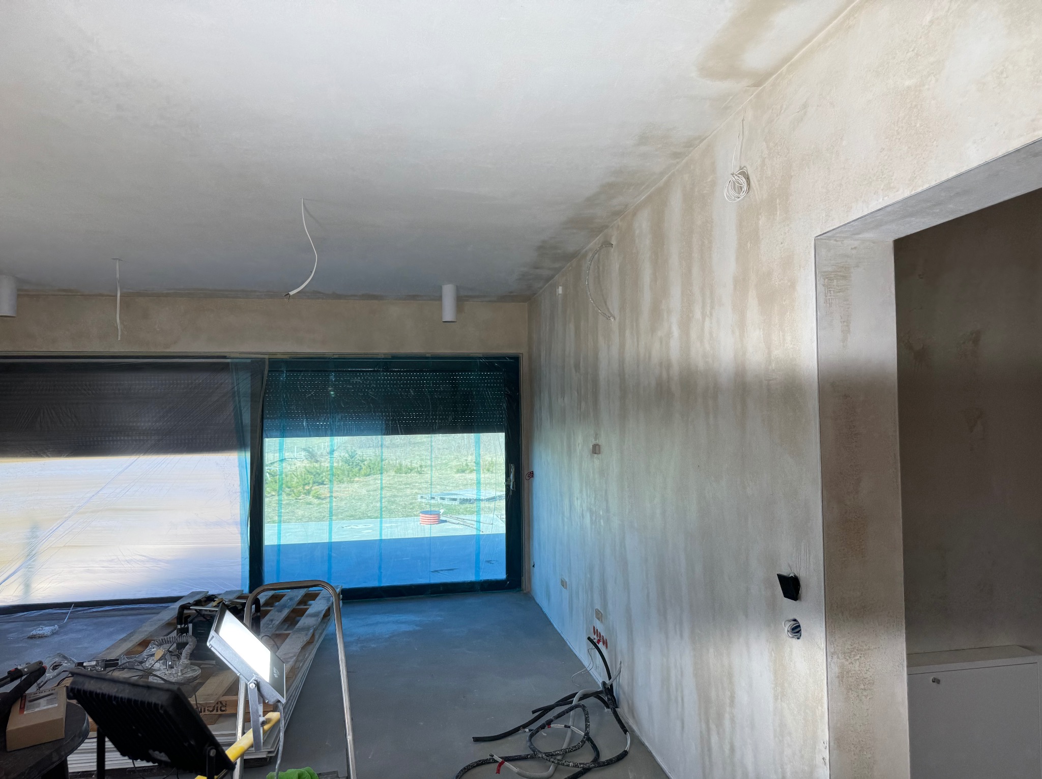 Wnętrze w trakcie prac wykończeniowych: ściany po szpachlowaniu, widoczne kable elektryczne, okno z roletą. Na podłodze narzędzia i lampa budowlana.
