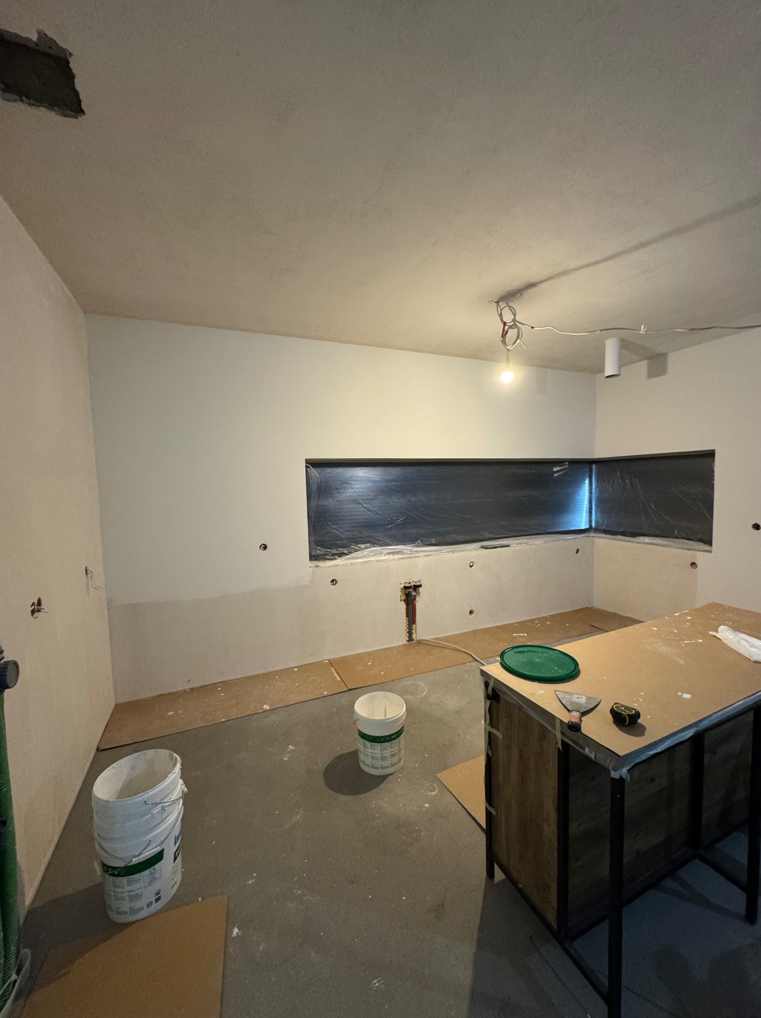 Wnętrze w trakcie prac wykończeniowych. Ściany częściowo wyszpachlowane, okno zabezpieczone folią, widoczne narzędzia i wiadro z farbą. Stan surowy, brak mebli.