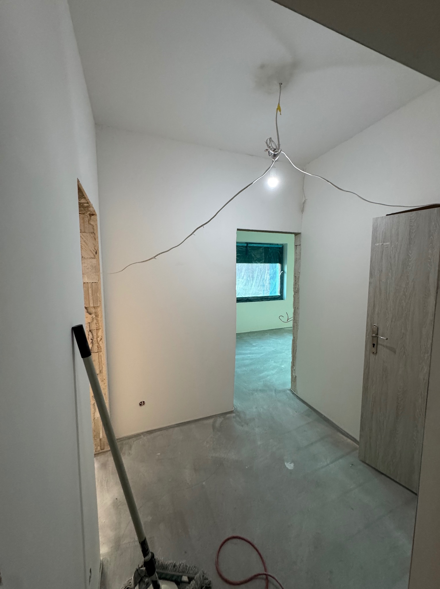 Wnętrze po szpachlowaniu i malowaniu ścian na biało, widoczny mop i kable wystające z sufitu, przygotowane do dalszych prac wykończeniowych.