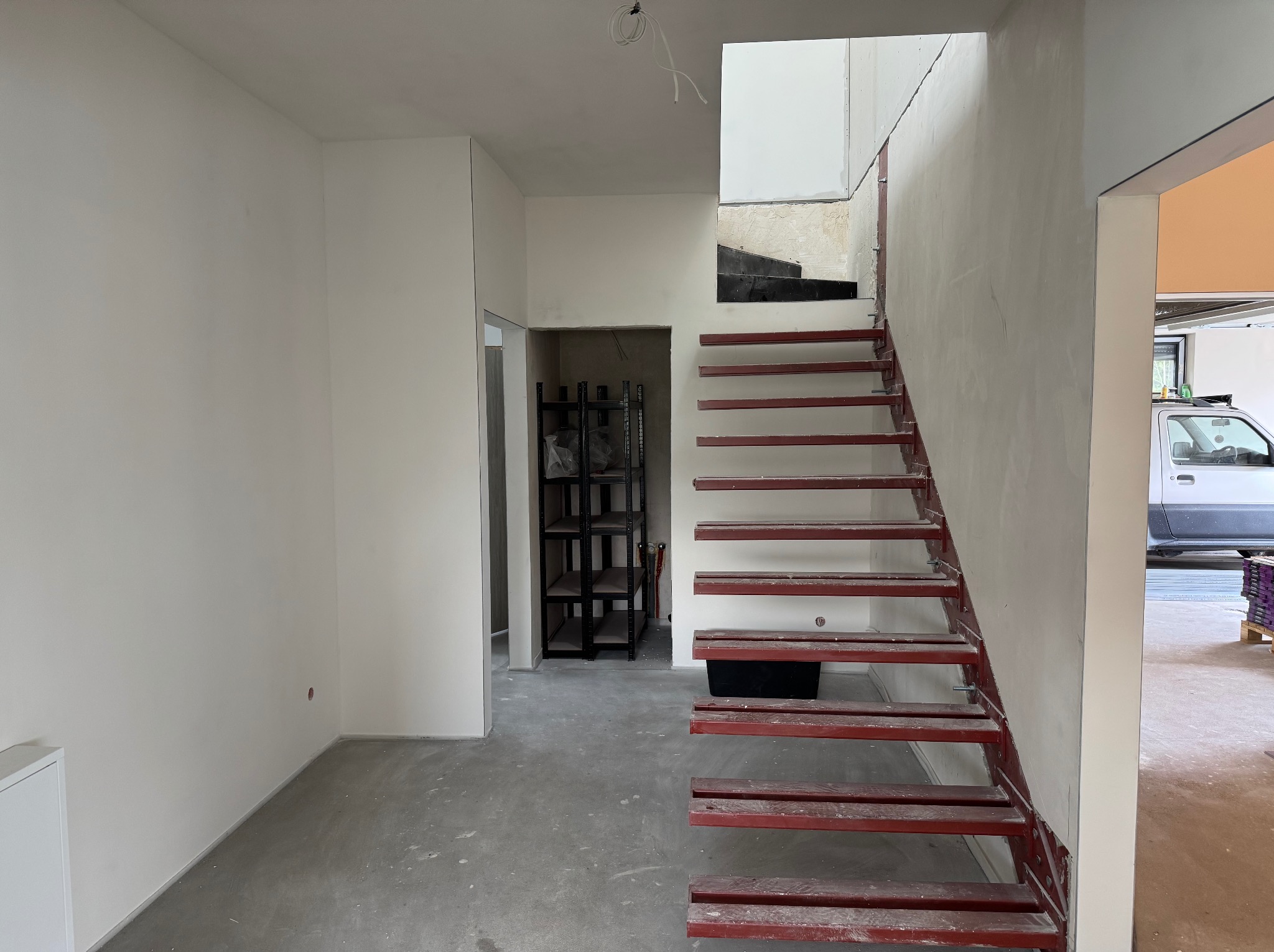Wnętrze w trakcie wykańczania: surowe schody stalowe, ściany po szpachlowaniu, widoczny regał w przejściu. Częściowo otynkowane ściany, instalacja elektryczna.