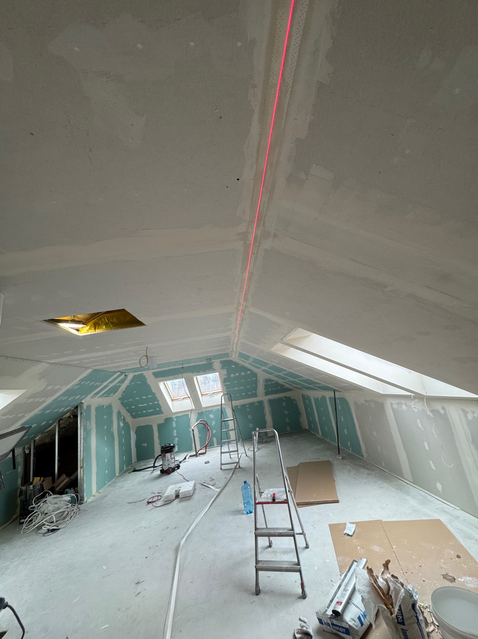 Montaż płyt karton-gips na poddaszu w Rumi, z użyciem poziomicy laserowej. Widoczne okna dachowe i surowe wykończenie.