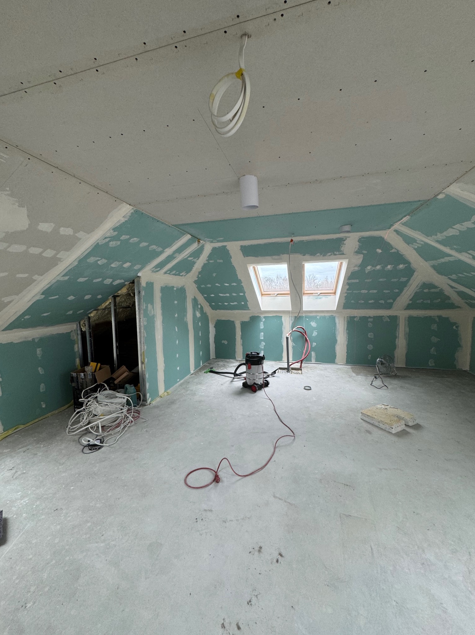 Poddasze w trakcie adaptacji: płyty karton-gips na ścianach i skosach, widoczne łączenia, okna dachowe, przewody elektryczne i narzędzia. Etap przygotowania do dalszych prac.