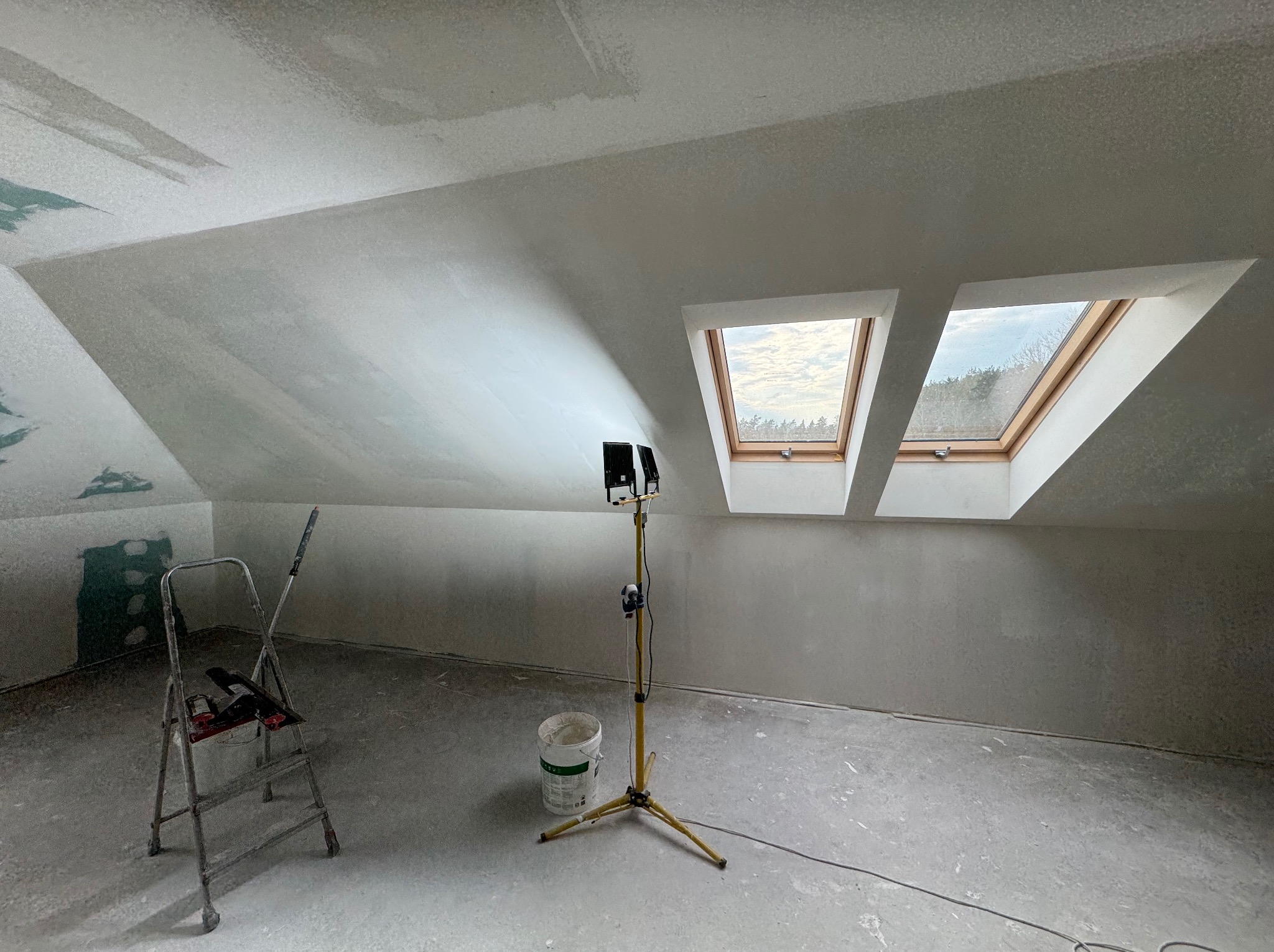 Poddasze po szpachlowaniu, widoczne okna dachowe, drabina i lampa budowlana. Ściany przygotowane do malowania, surowe wykończenie.