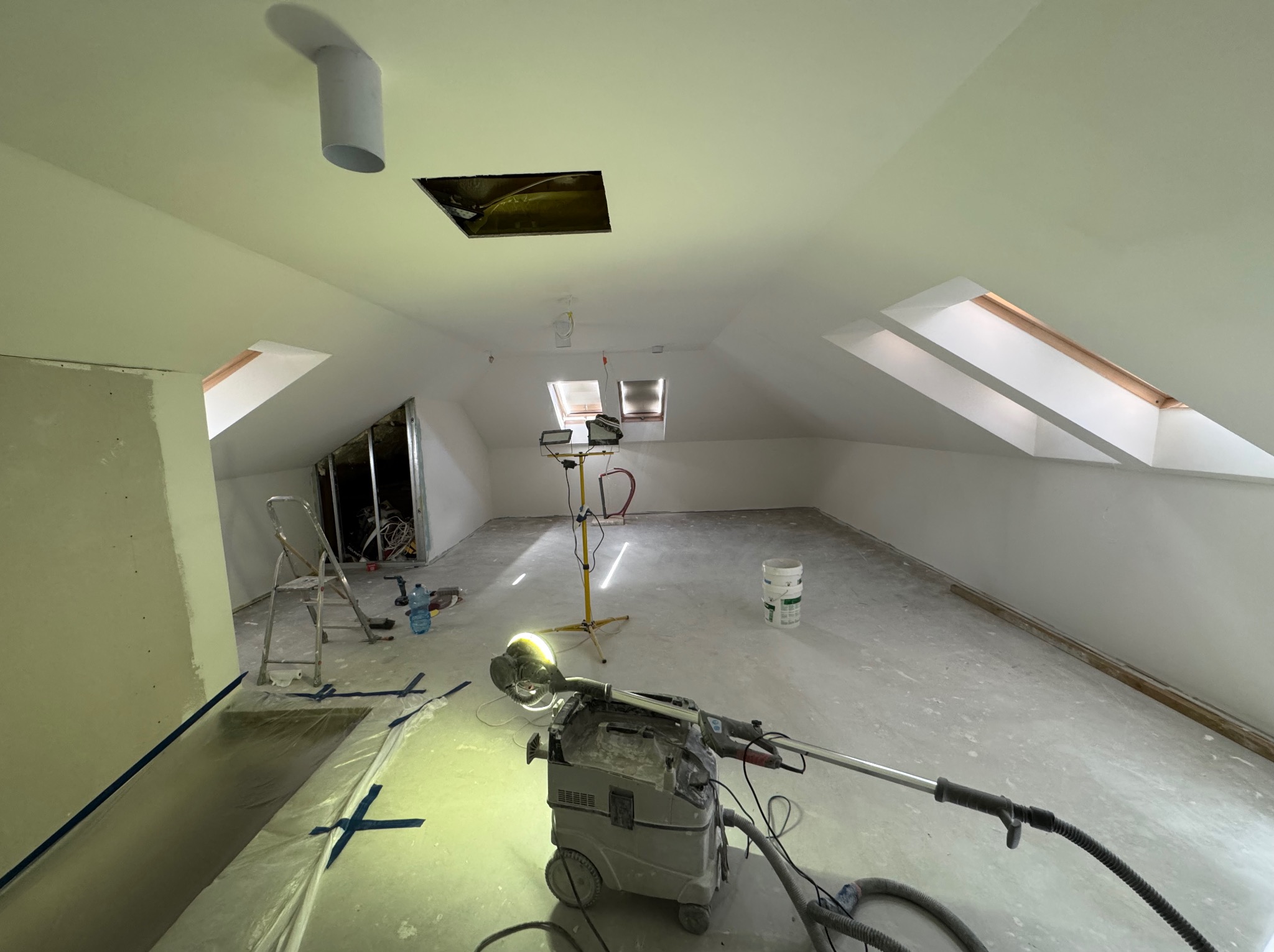 Poddasze w trakcie szpachlowania z widoczną szlifierką do gipsu, zabezpieczoną folią podłogą i oknami dachowymi. Widoczne narzędzia i lampa oświetleniowa.