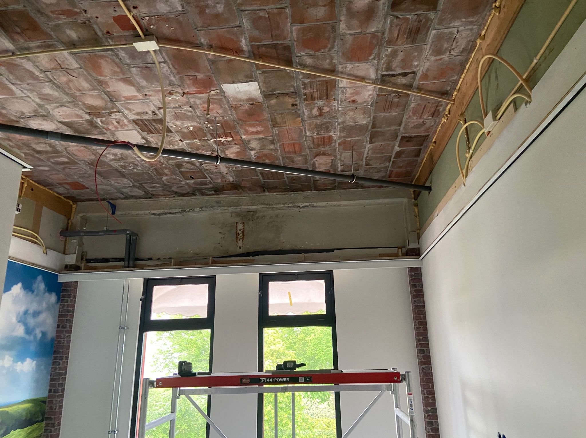 Wnętrze w trakcie remontu: surowy ceglany sufit z instalacjami, świeżo pomalowane ściany i rusztowanie z poziomnicą. Widok na zieleń przez okna.