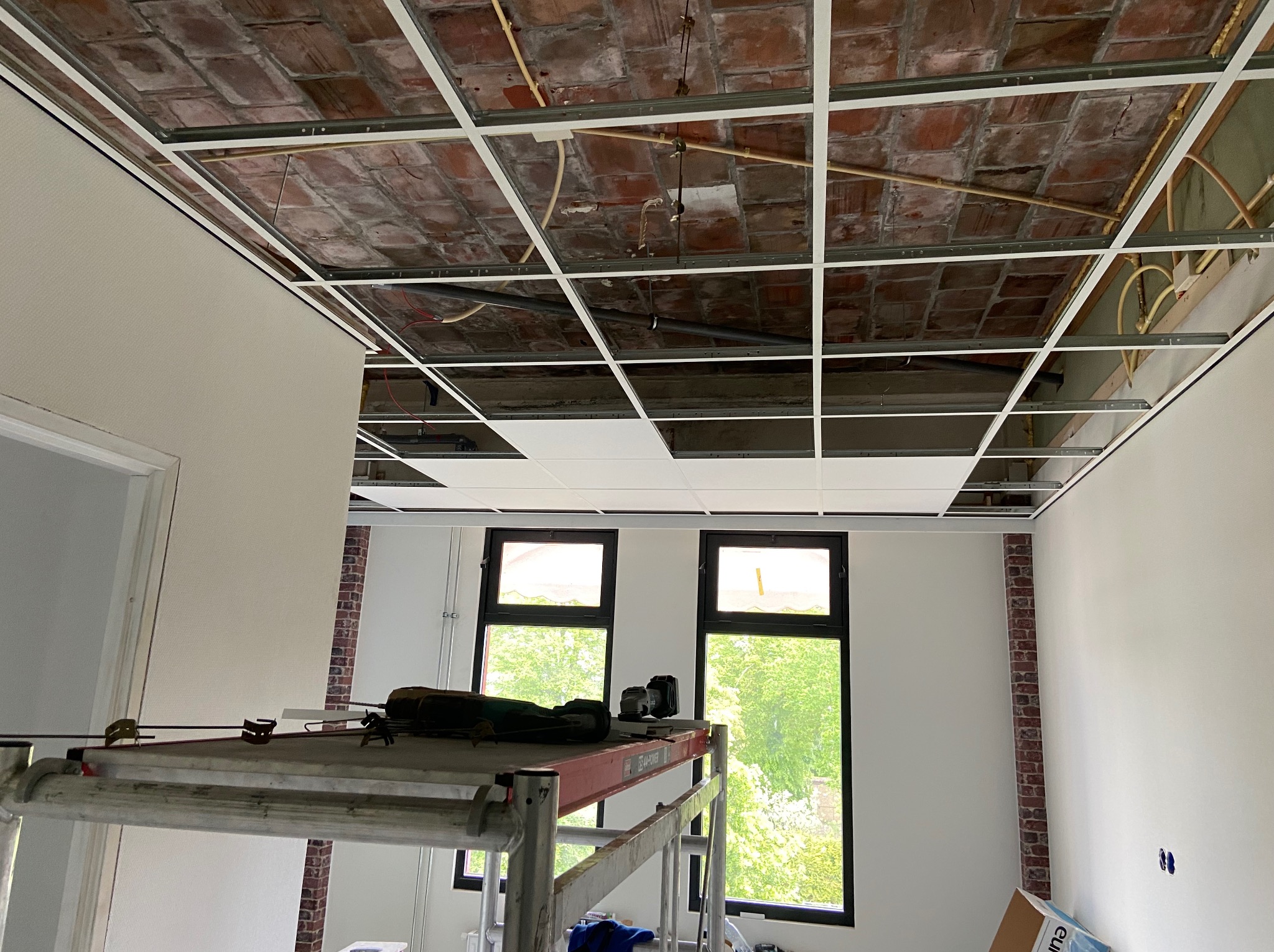 Montaż sufitu podwieszanego z widoczną konstrukcją metalową i częściowo włożonymi panelami. W tle okna z zielenią i rusztowanie.