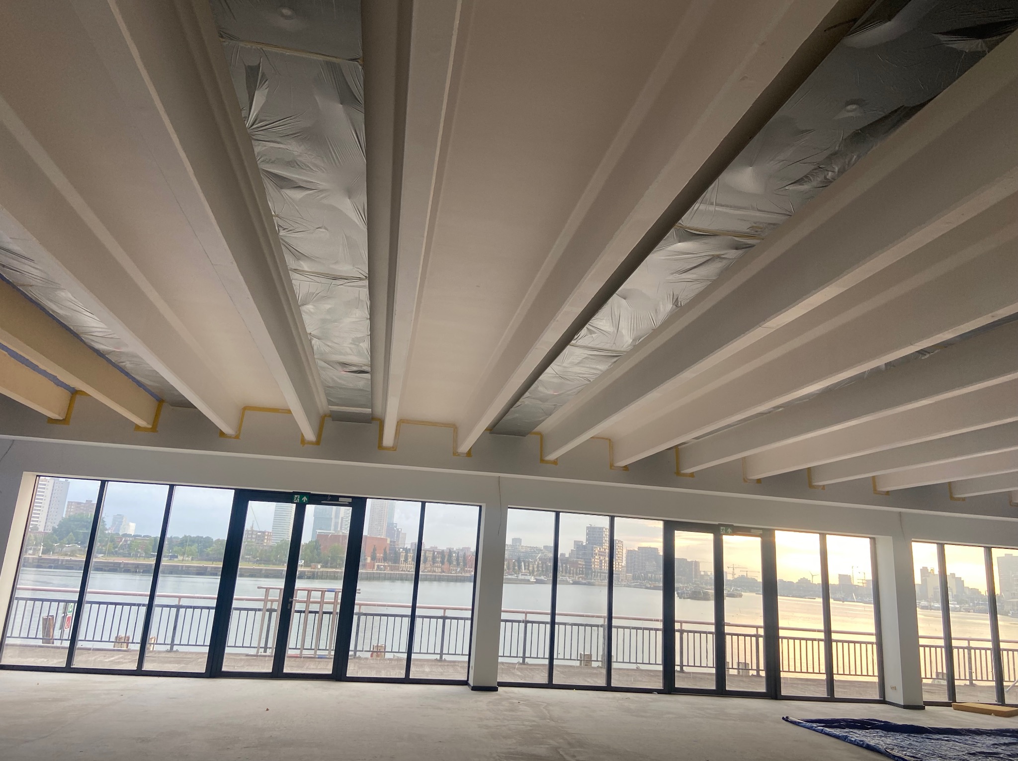 Wnętrze w trakcie wykańczania z widokiem na miasto przez duże okna. Sufit z belkami zabezpieczonymi folią i taśmą malarską. Podłoga przygotowana do dalszych prac.