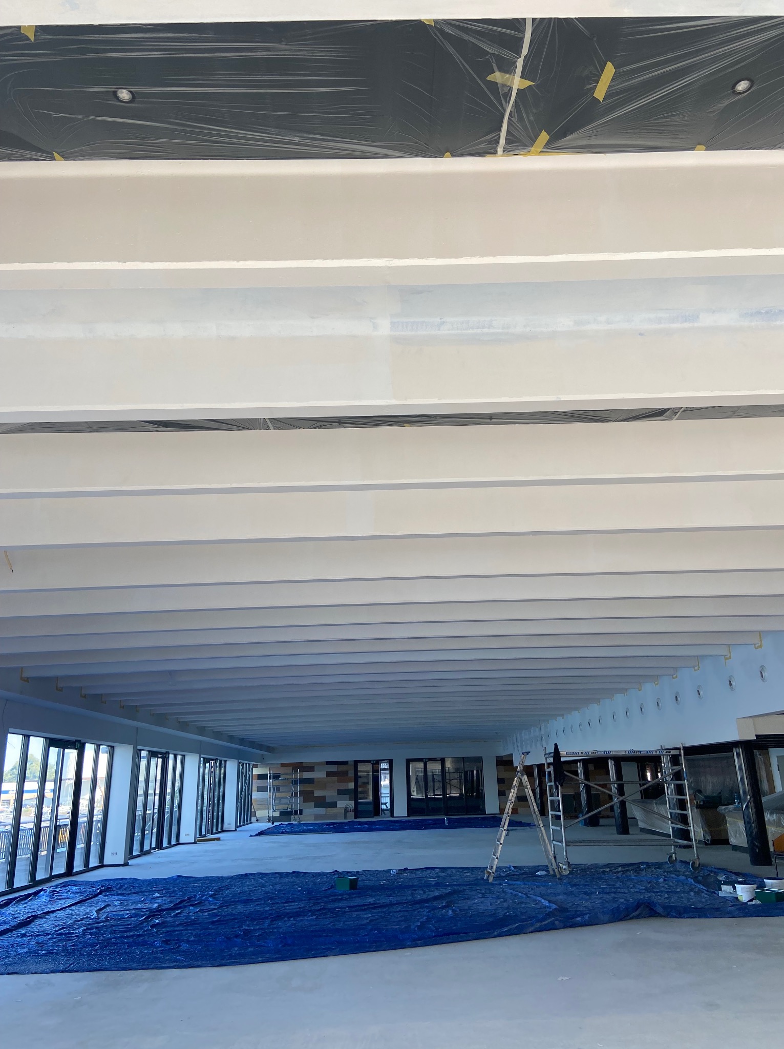 Wnętrze komercyjne w trakcie malowania: białe belki stropowe, zabezpieczona folią sufitowa, podłoga przykryta niebieską folią ochronną, widoczne rusztowania i okna.