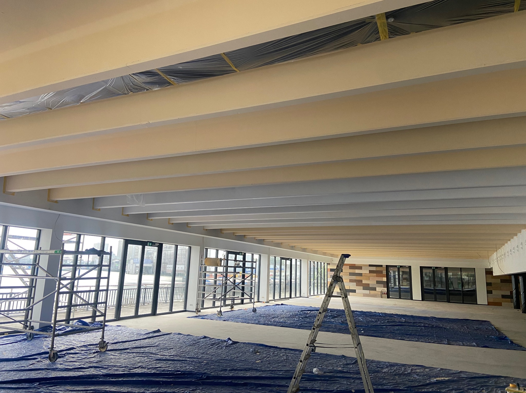 Wnętrze w trakcie malowania: jasne belki sufitowe, rusztowania i drabina. Podłoga zabezpieczona folią malarską. Duże okna z widokiem na zewnątrz.