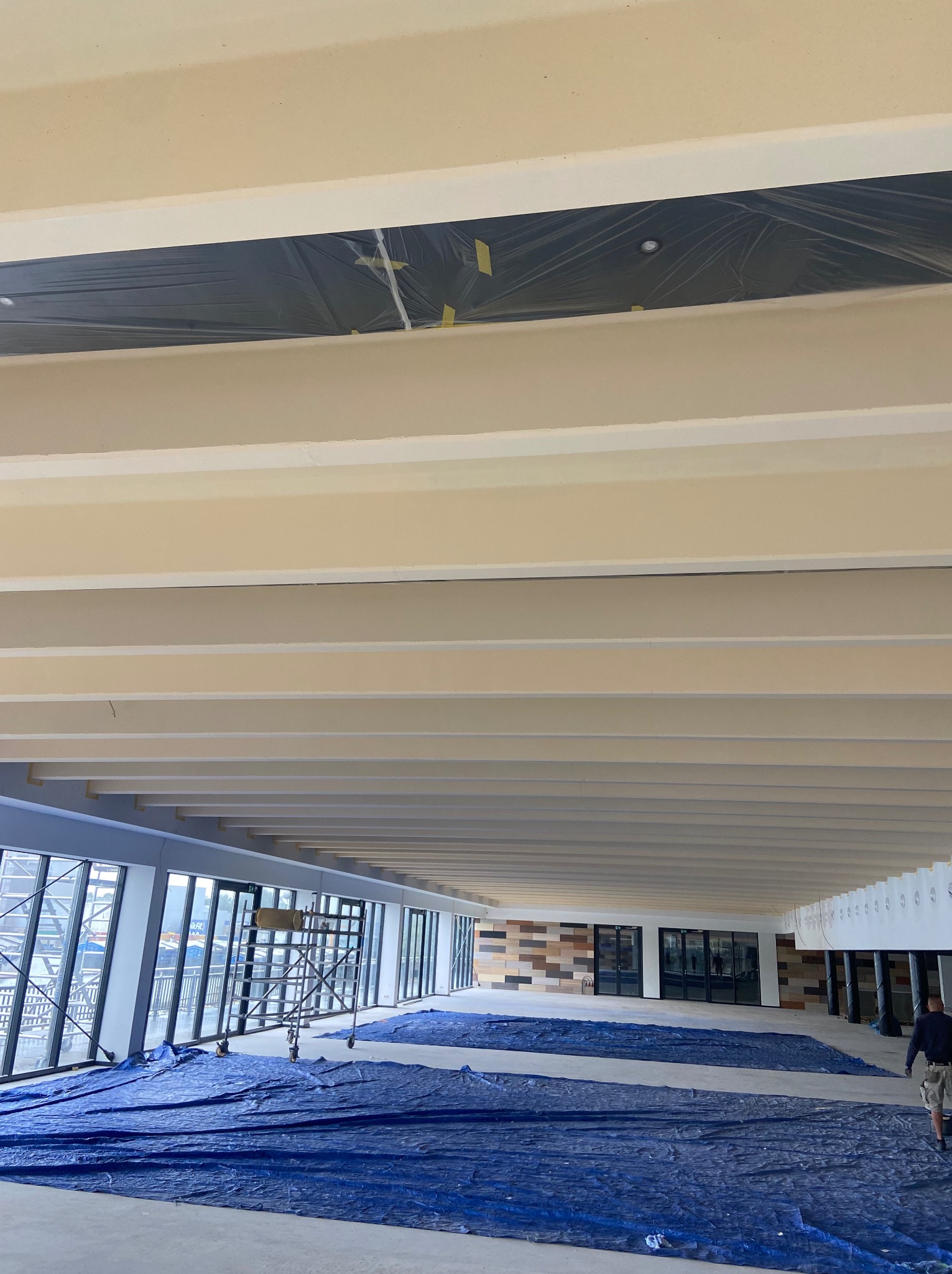 Wnętrze w trakcie prac wykończeniowych: beżowe belki stropowe, okna z widokiem na zewnątrz, podłoga zabezpieczona folią, rusztowanie i fragment sylwetki osoby.