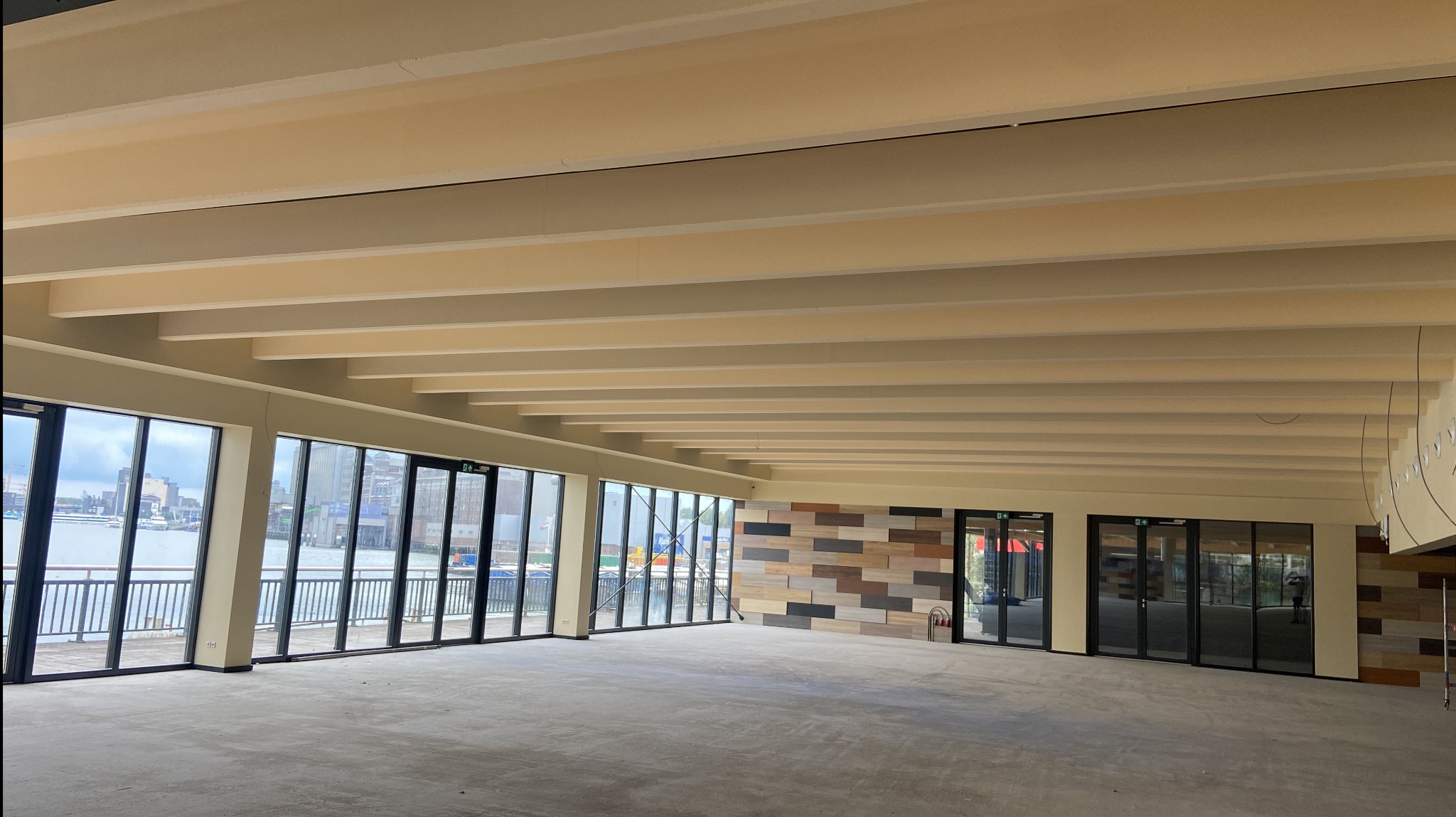 Przestronne, jasne wnętrze w trakcie wykańczania, z dużymi oknami z widokiem na wodę i dekoracyjną ścianą z paneli w odcieniach brązu i beżu. Widoczne beżowe belki stropowe.