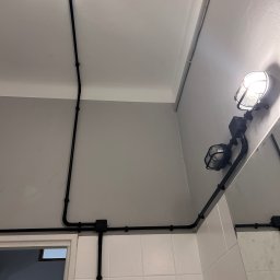 ELECTROFIBER PRZEMYSŁAW SAWICKI - Dwa reflektory przemysłowe zamontowane na szarej ścianie z widocznymi czarnymi przewodami elektrycznymi, tworzącymi instalację w stylu industrialnym.