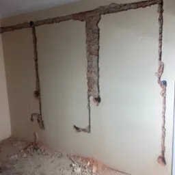 Ściana z wyżłobieniami pod instalację elektryczną, widoczne przewody i puszki. Fragmenty cegieł i gruzu na podłodze. Prace przygotowawcze do instalacji.