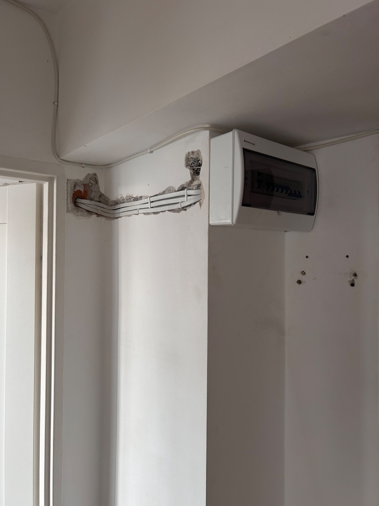 Biała skrzynka rozdzielcza z widocznymi kablami w ścianie, świeżo po montażu. Widoczne ślady po kuciu w ścianie. Kable ułożone w korytku.