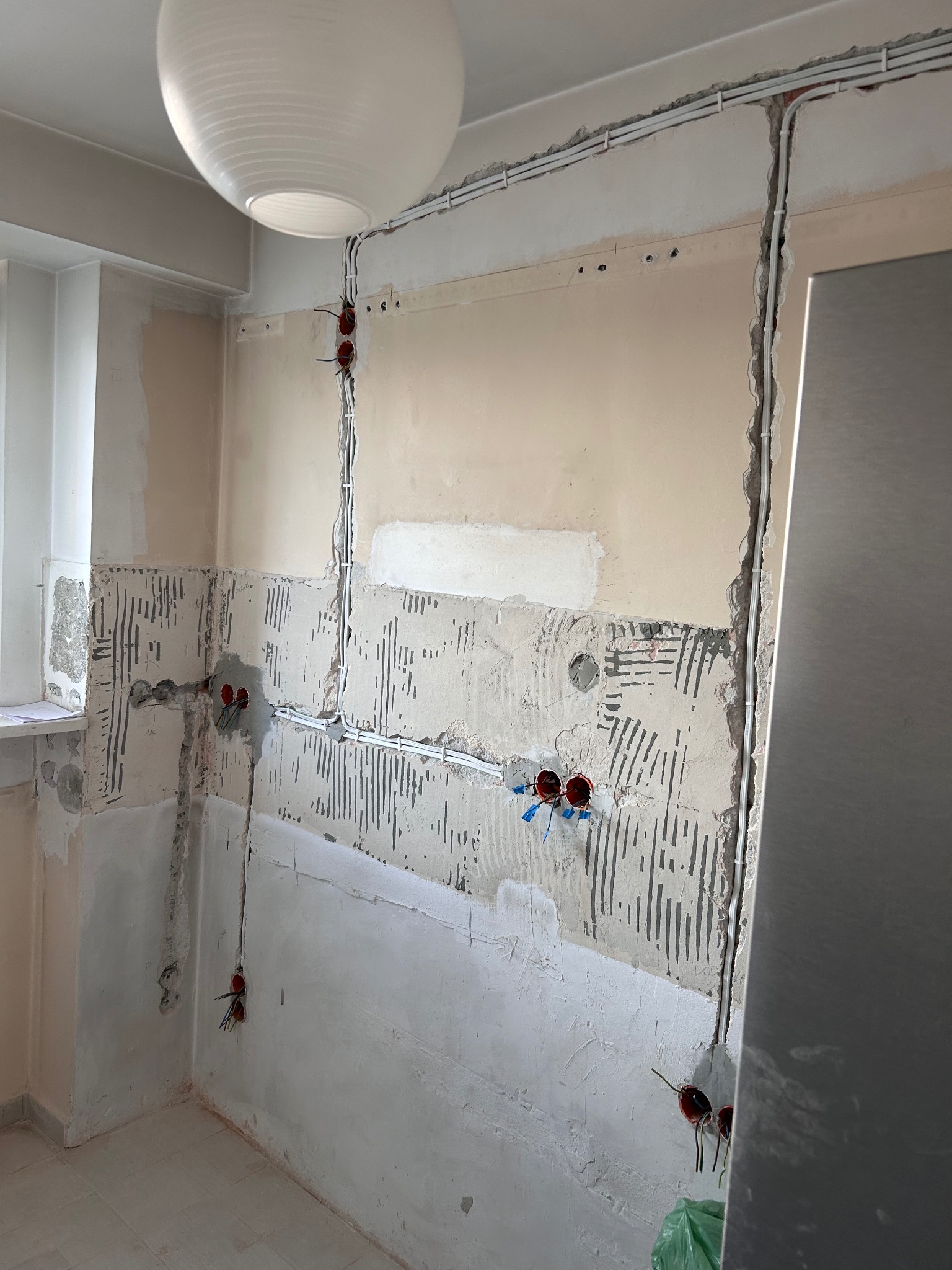 Ściana w trakcie remontu z widocznymi przewodami elektrycznymi w bruzdach, puszkami instalacyjnymi. Widoczny fragment okna i wisząca lampa.