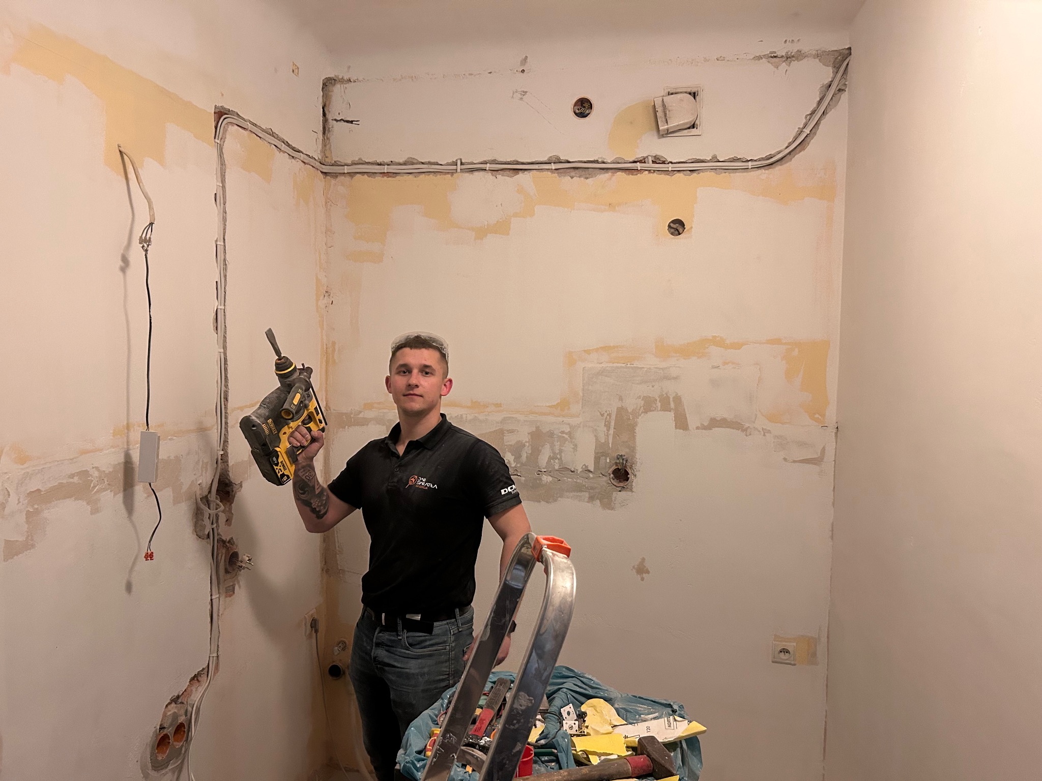 Elektryk z wiertarką udarową na tle ściany z instalacją elektryczną w trakcie montażu. Widoczne kable, puszki i narzędzia.