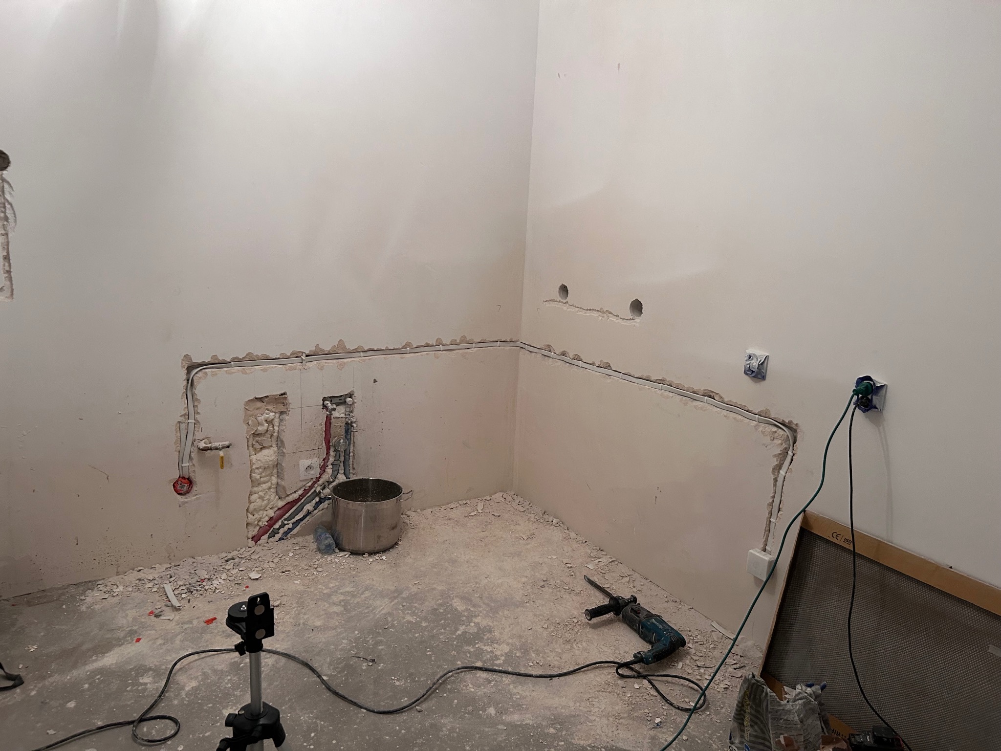 Remont kuchni w trakcie: wyfrezowane bruzdy pod kable elektryczne i rury, widoczny sprzęt (wiertarka, poziomica na statywie), gruz na podłodze. Stan surowy.
