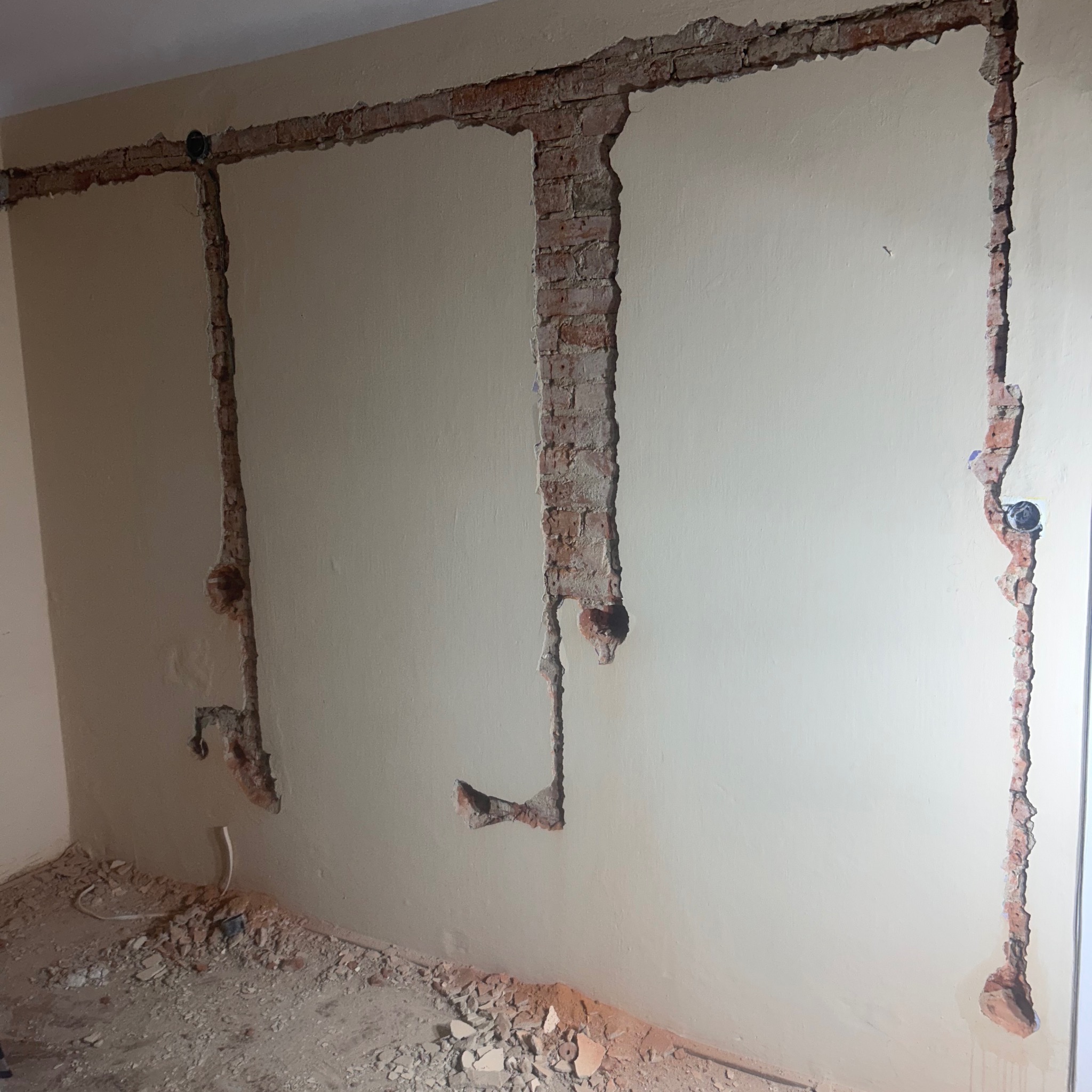 Ściana z wyżłobieniami pod instalację elektryczną, widoczne przewody i puszki. Fragmenty cegieł i gruzu na podłodze. Prace przygotowawcze do instalacji.