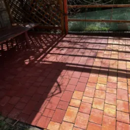 Czerwono-pomarańczowa kostka brukowa pod zadaszeniem z drewnianą konstrukcją, częściowo zacieniona, z widocznym fragmentem zieleni.