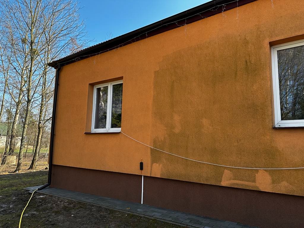 Czyszczenie elewacji budynku z widoczną różnicą w kolorze ścian po umyciu. W tle drzewa bez liści i fragment nieba.
