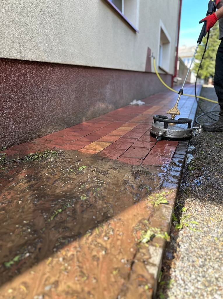 Czyszczenie kostki brukowej z użyciem myjki ciśnieniowej. Widoczny fragment chodnika przed i po zabiegu. Woda i resztki zanieczyszczeń na powierzchni kostki.