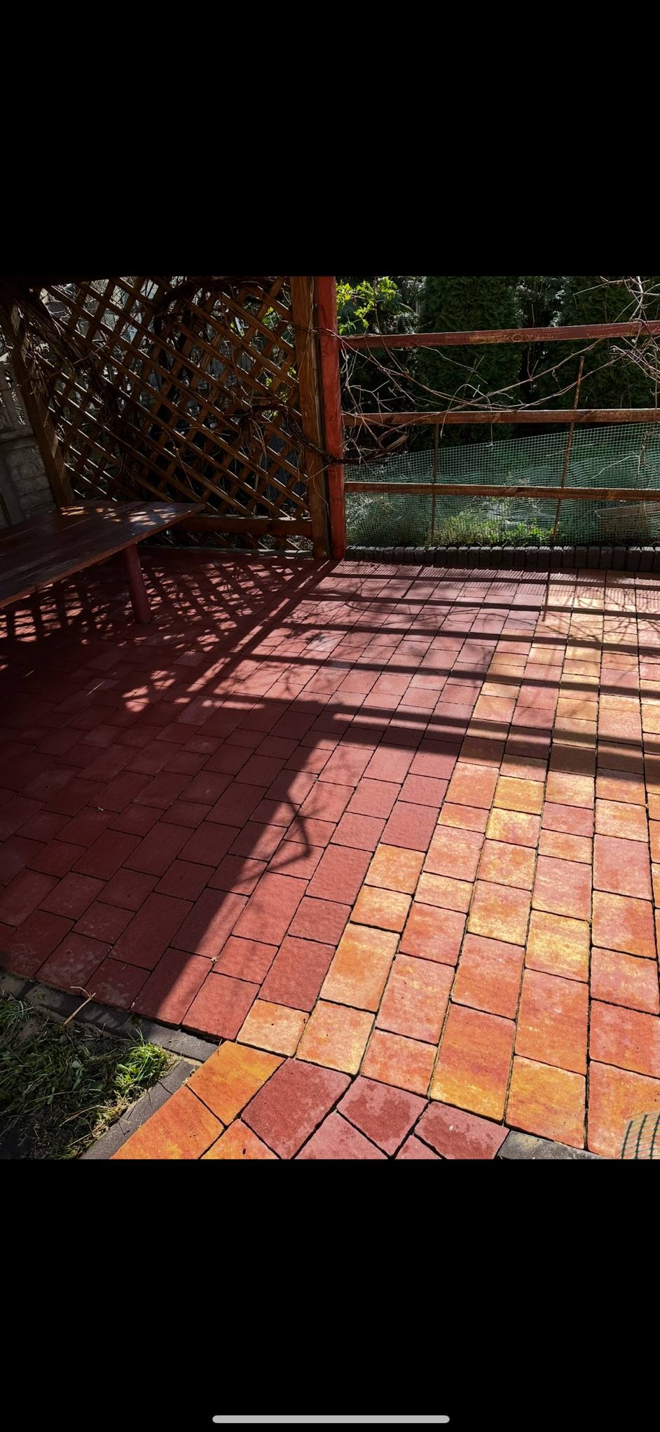 Czerwono-pomarańczowa kostka brukowa pod zadaszeniem z drewnianą konstrukcją, częściowo zacieniona, z widocznym fragmentem zieleni.