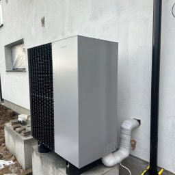 Pogotowie Hydrauliczne - Szara pompa ciepła Viessmann zamontowana na betonowych fundamentach przy białej ścianie budynku, widoczny czarny spust wody deszczowej i rura odprowadzająca skropliny owinięta taśmą.