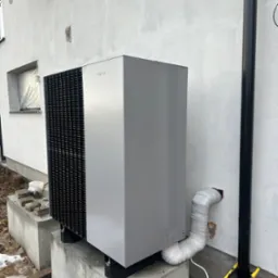 Szara pompa ciepła Viessmann zamontowana na betonowych fundamentach przy białej ścianie budynku, widoczny czarny spust wody deszczowej i rura odprowadzająca skropliny owinięta taśmą.