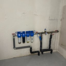 Pogotowie Hydrauliczne - Zamontowany system filtrów wody z czterema cylindrami i manometrami, rury z izolacją termiczną, szare płytki podłogowe, surowa ściana. Instalacja wodna w pomieszczeniu.