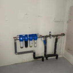 Zamontowany system filtrów wody z czterema cylindrami i manometrami, rury z izolacją termiczną, szare płytki podłogowe, surowa ściana. Instalacja wodna w pomieszczeniu.