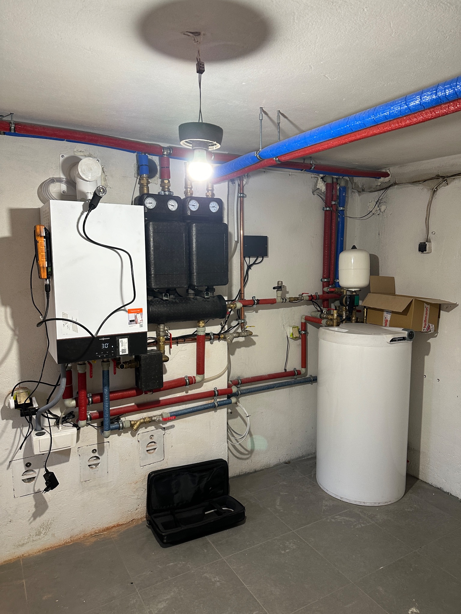 Nowoczesny piec gazowy Viessmann z czarnym osprzętem i białym zasobnikiem ciepłej wody, połączone rurami w kolorach niebieskim i czerwonym. Na podłodze leży czarna torba narzędziowa.