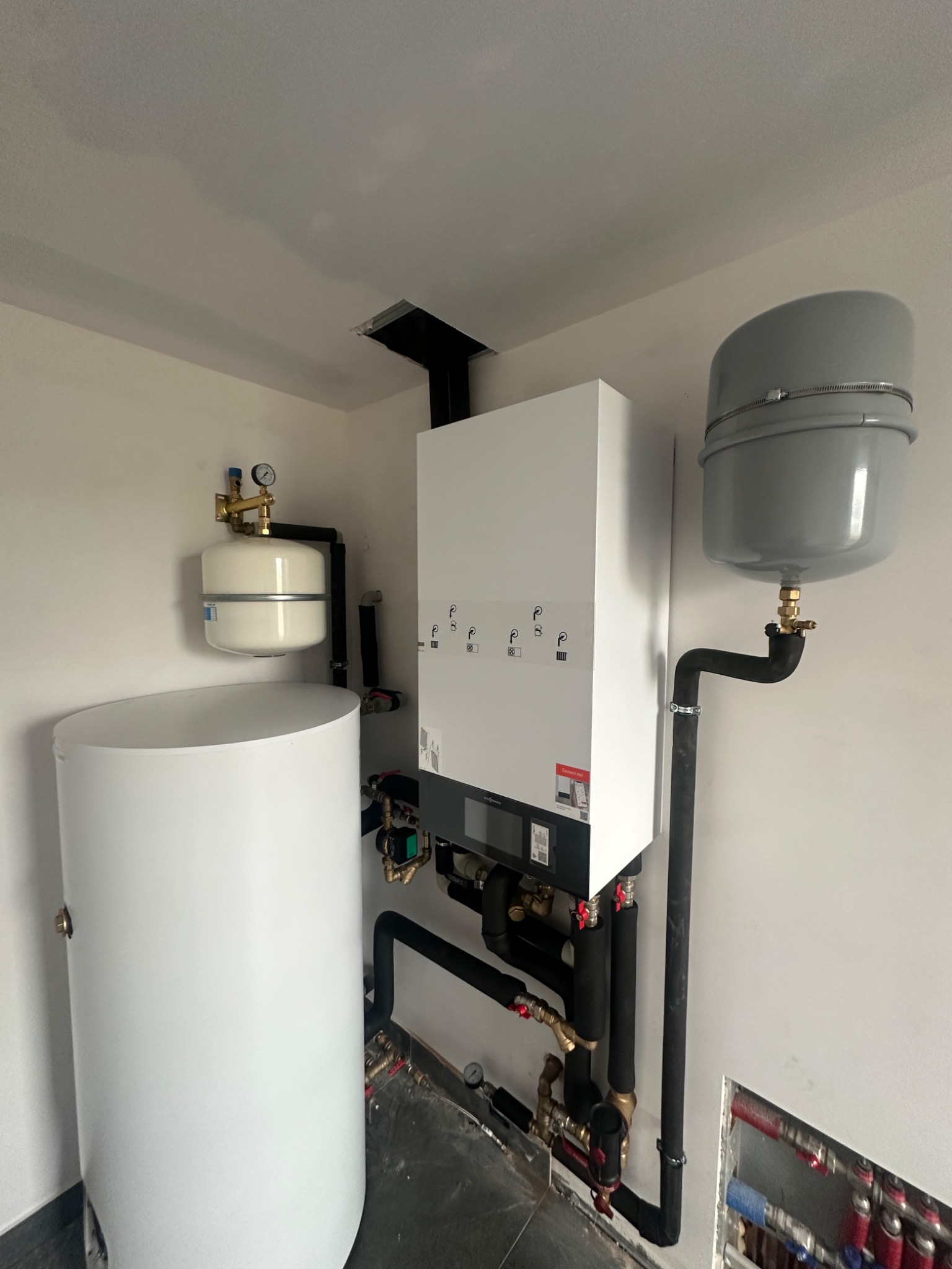 Nowoczesny, biały piec gazowy Viessmann zamontowany na ścianie, z widocznymi czarnymi rurami instalacji, białym i szarym zasobnikiem oraz fragmentem ogrzewania podłogowego w ścianie, w surowym...