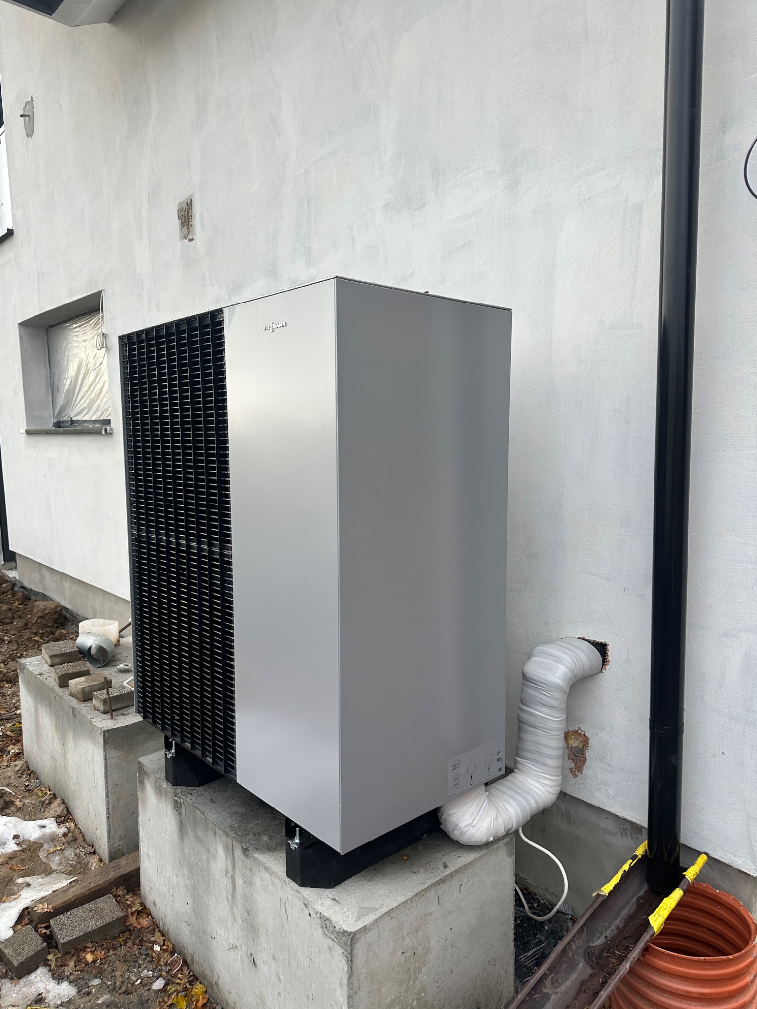 Szara pompa ciepła Viessmann zamontowana na betonowych fundamentach przy białej ścianie budynku, widoczny czarny spust wody deszczowej i rura odprowadzająca skropliny owinięta taśmą.