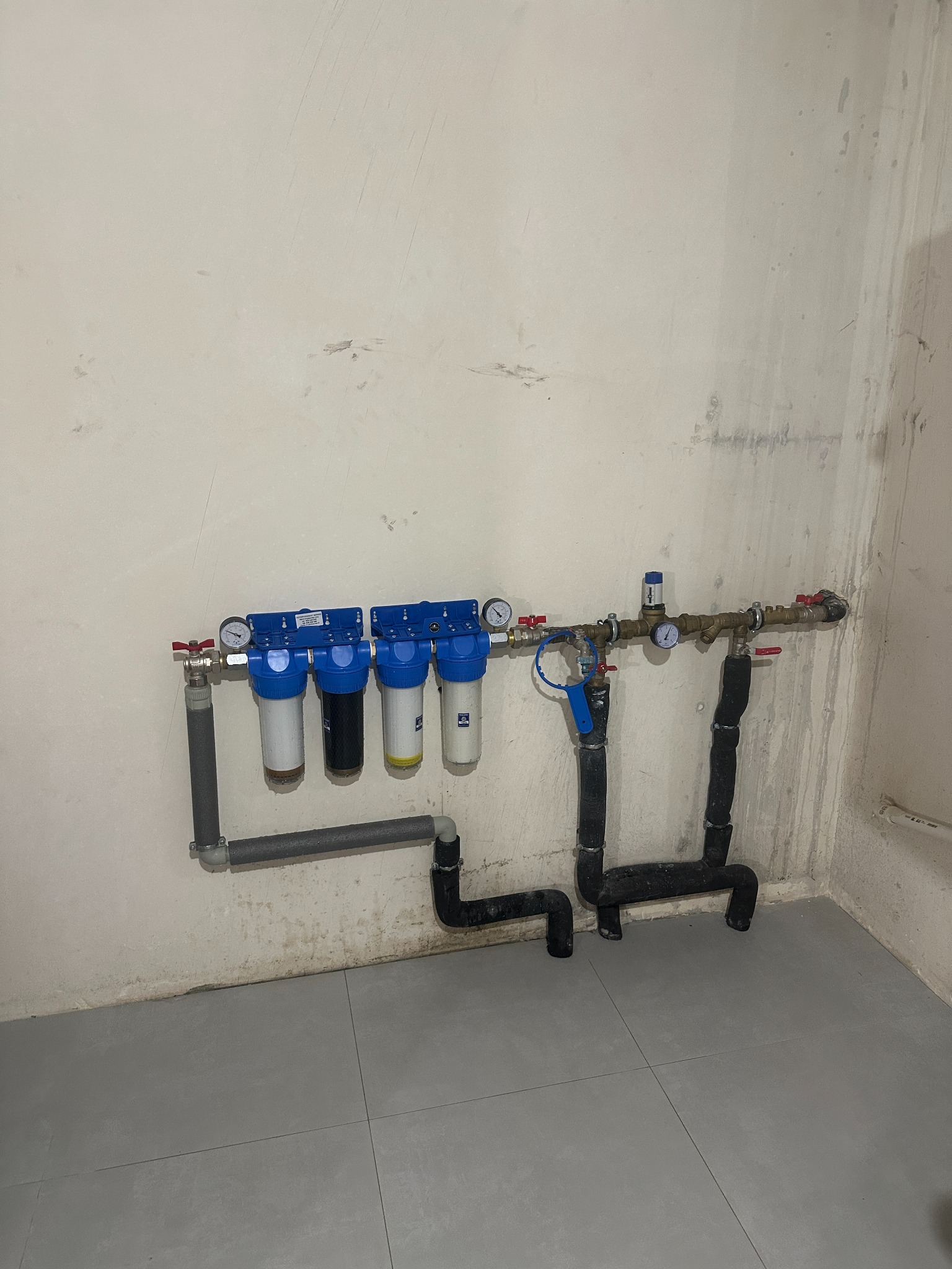 Zamontowany system filtrów wody z czterema cylindrami i manometrami, rury z izolacją termiczną, szare płytki podłogowe, surowa ściana. Instalacja wodna w pomieszczeniu.