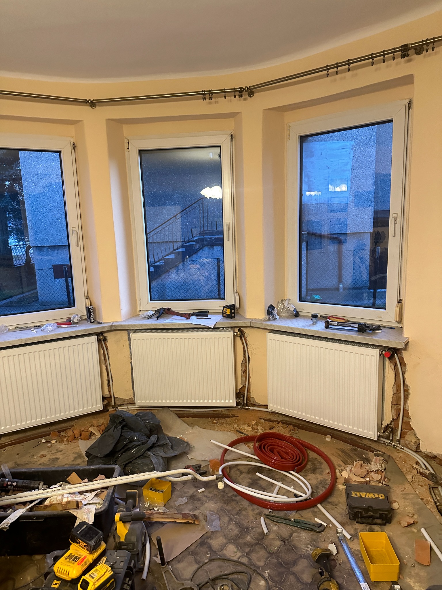 Remont pokoju z trzema oknami i grzejnikami. Widoczne narzędzia, rury i gruz. W tle zabudowa okienna z karniszem.