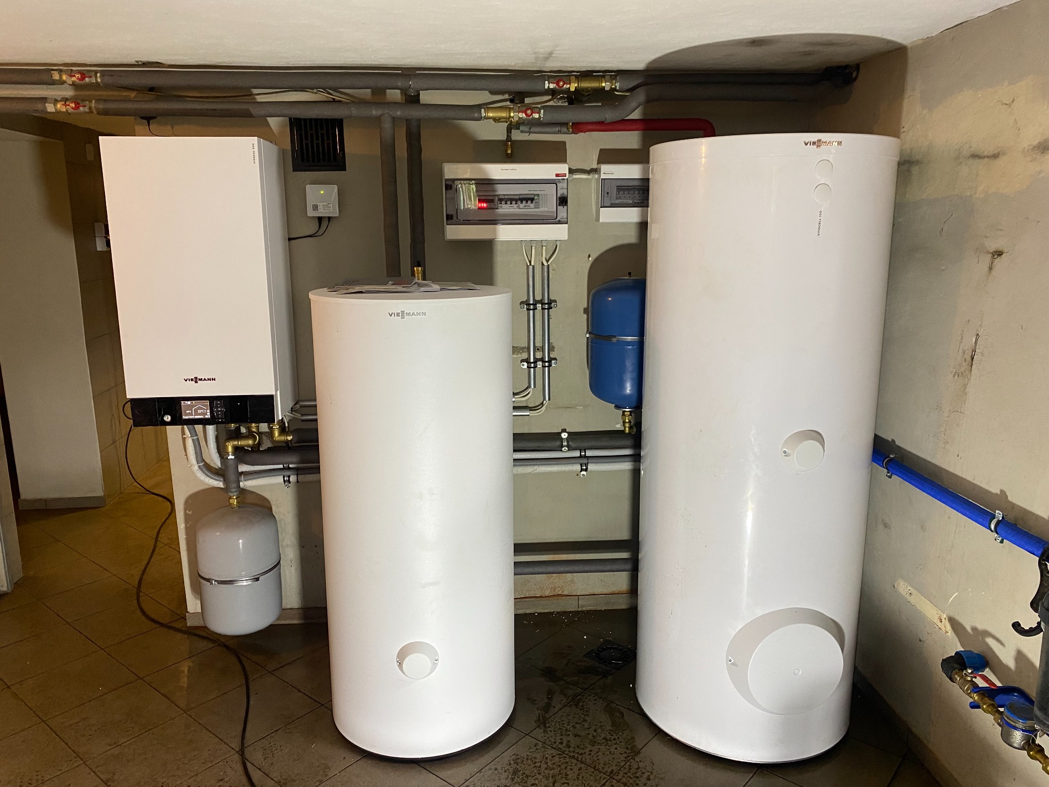Biały piec gazowy Viessmann oraz dwa białe zasobniki ciepłej wody w piwnicy, widoczne rury instalacji grzewczej i elektryczna skrzynka bezpiecznikowa na ścianie.