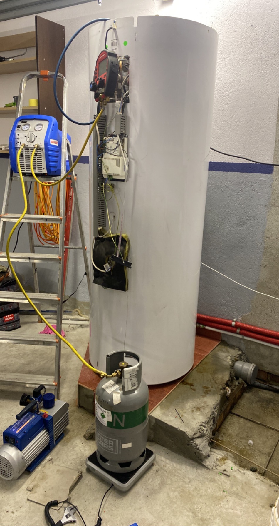 Biały piec gazowy w trakcie montażu z podłączonymi narzędziami, butla gazowa obok. Widoczna drabina i ściana w tle. Prace instalacyjne w pomieszczeniu.