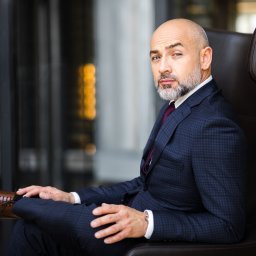 Mężczyzna z brodą w garniturze w kratę siedzi w skórzanym fotelu, spoglądając w obiektyw. Elegancki strój, brązowe buty, stonowane tło. Sesja biznesowa.