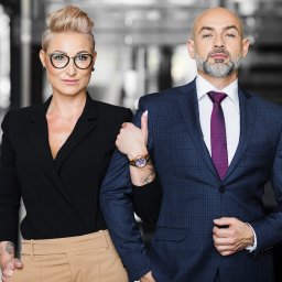 Profesjonalny portret biznesowy w Łodzi: elegancka kobieta w okularach opiera rękę na ramieniu mężczyzny w garniturze. Stonowane tło biurowe, oświetlenie studyjne.
