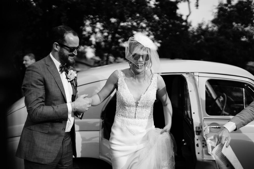 Uśmiechnięta panna młoda w koronkowej sukni wychodzi z zabytkowego auta, trzymając za rękę pana młodego w garniturze. Czarno-biała fotografia ślubna.