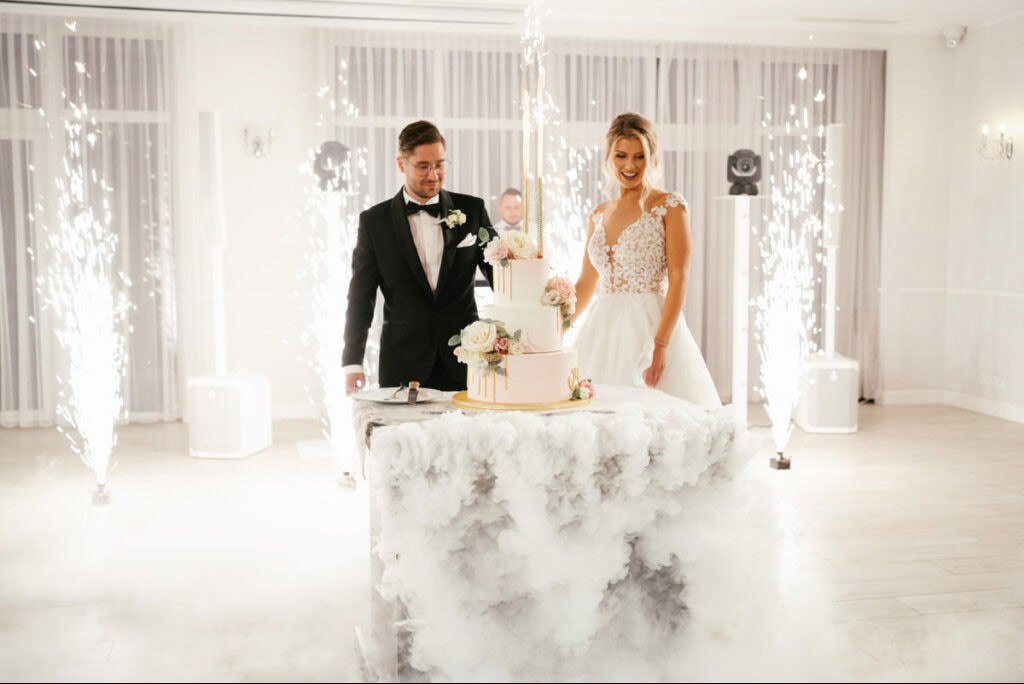Uśmiechnięta para młoda kroi tort weselny, otoczona fontannami iskier i efektem dymu. Eleganckie wnętrze sali balowej z białymi zasłonami w tle.